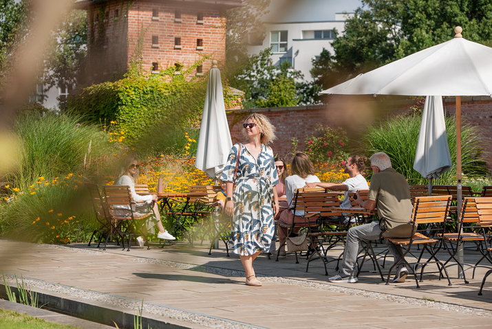 Eine Frau läuft im Sommerkleid durch den Englischen Garten, dahinter sitzen Menschen an Cafétischen